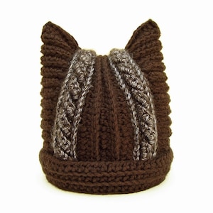 Könnte beinhalten: Braune Strickmütze mit Katzenohren. Die Mütze hat ein braunes und graues Zopfmuster.