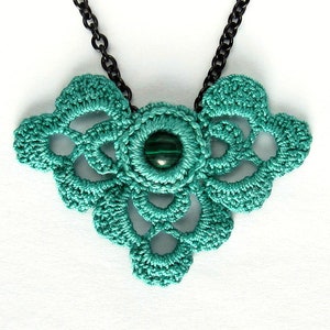 May include: A handmade teal crochet necklace with a central green stone. The necklace features an intricate openwork design and hangs from a black chain. The pendant has a floral shape, showcasing the delicate craftsmanship.
