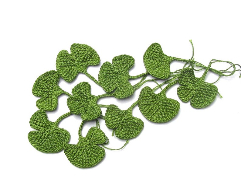 K&ouml;nnte beinhalten: Ein Set aus zehn gr&uuml;nen geh&auml;kelten Ginkgobl&auml;ttern, jedes mit einem zarten, f&auml;cherf&ouml;rmigen Design. Die Bl&auml;tter sind durch d&uuml;nne F&auml;den verbunden, was einen flie&szlig;enden, organischen Look erzeugt.