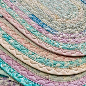 May include: Close-up of several oval-shaped, quilted mats in pastel colors. The mats are layered, showcasing a variety of hues including blue, pink, and green. Each mat has a scalloped edge and a quilted pattern, creating a textured appearance.
