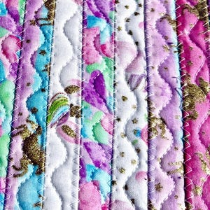 May include: Close-up of a quilted fabric with a floral pattern in shades of pink, purple, blue, and green. The fabric has a white wavy stitched pattern and gold accents. The quilting is done with a zigzag stitch.