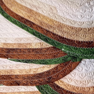 May include: Close-up of a quilted textile with a repeating pattern of curved, layered bands. The bands feature shades of white, tan, brown, and green, with decorative stitching. The quilt has a handmade aesthetic.