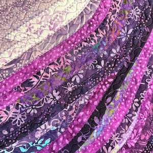 May include: Close-up of a quilted textile with a vibrant array of purple hues. The design features curved, layered strips of fabric with various patterns, including floral and dotted motifs, stitched together with contrasting thread.