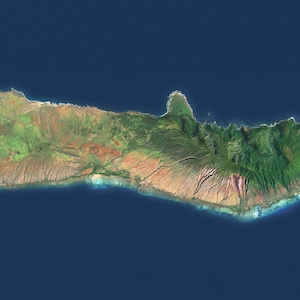 May include: Satellite view of a long, narrow island surrounded by deep blue ocean. The island features a diverse landscape with green, brown, and red hues, suggesting varied vegetation and terrain. Coastal areas show a lighter blue, indicating shallow waters.