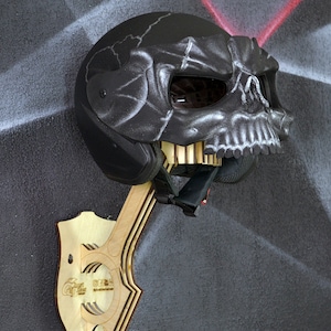 Pode incluir: Um capacete de moto preto com um design de caveira está montado em um suporte de parede de madeira. O suporte é feito de madeira em camadas e tem um design de corte. O capacete tem um design de caveira preto e branco com um contorno branco.