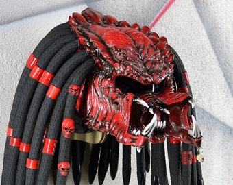 May include: A red and black motorcycle helmet with a predator-like design and dreadlocks. The helmet has a black visor and red accents.