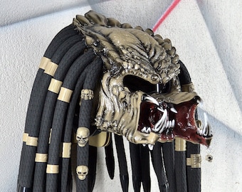 Könnte beinhalten: Ein detaillierter Predator-Helm mit einem strukturierten, beigefarbenen Gesicht und offenem Mund, der scharfe weiße Zähne und einen roten Innenraum zeigt. Der Helm hat schwarze Dreadlocks mit beigen Akzenten und kleinen Totenkopfdekorationen. Das Design ist aufwendig und bedrohlich.
