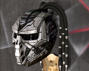 Puede incluir: Un casco de moto plateado y negro con un diseño futurista. El casco tiene una visera y una larga trenza negra con detalles plateados. El casco está montado en un soporte de madera.