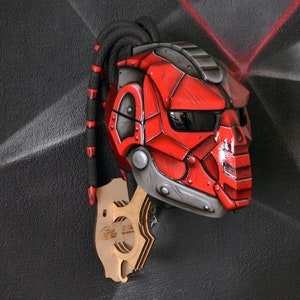 Puede incluir: Un casco rojo y gris con un diseño futurista, con una cara segmentada y una visera oscura. El casco tiene una cola trenzada negra y está montado en una pieza de exhibición de madera. El fondo es una pared gris texturizada.