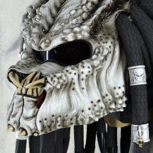 May include: A white Predator-style motorcycle helmet with black dreadlocks and a black visor. The helmet has a detailed design with sharp teeth and a textured surface.