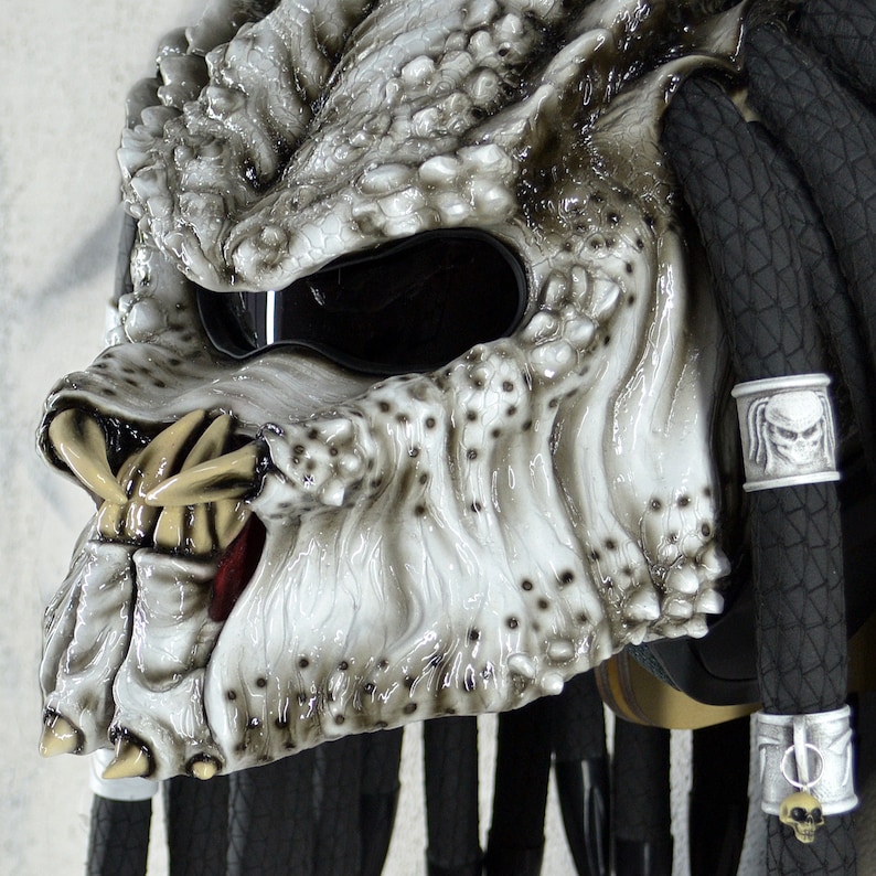 May include: A white Predator-style motorcycle helmet with black dreadlocks and a black visor. The helmet has a detailed design with sharp teeth and a textured surface.
