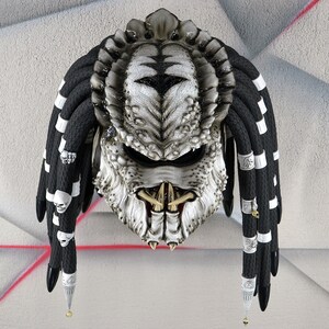 May include: A white and black motorcycle helmet with dreadlocks. The helmet is designed to look like a Predator from the movie franchise. The helmet has a black visor and a white skull design on the front.