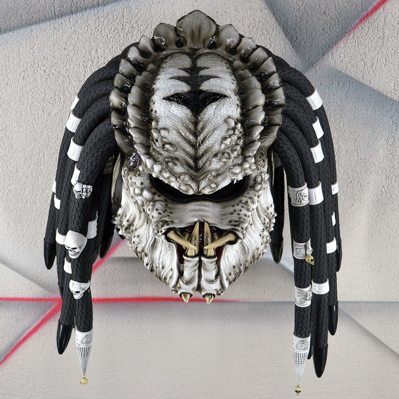 May include: A white and black motorcycle helmet with dreadlocks. The helmet is designed to look like a Predator from the movie franchise. The helmet has a black visor and a white skull design on the front.