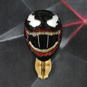 May include: A black motorcycle helmet with a white and red Venom design. The helmet has a large, toothy grin and is mounted on a wooden stand.