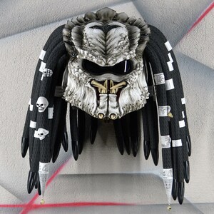 May include: A white and silver Predator-style motorcycle helmet with black dreadlocks. The helmet has a visor and a detailed design.