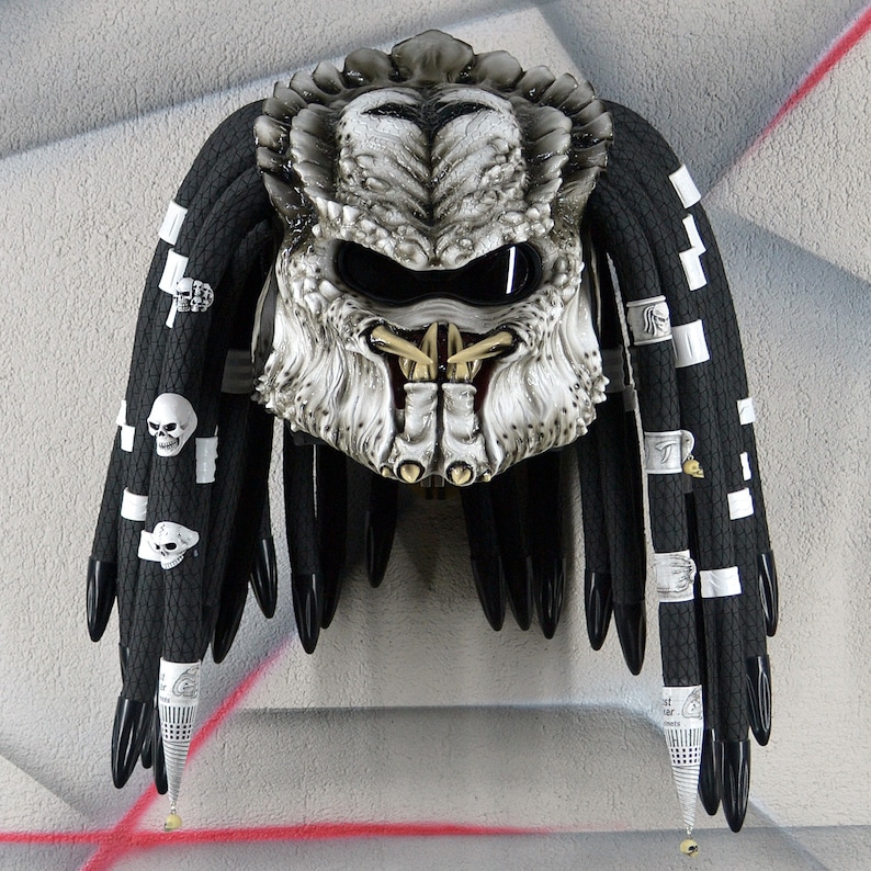 May include: A white and silver Predator-style motorcycle helmet with black dreadlocks. The helmet has a visor and a detailed design.
