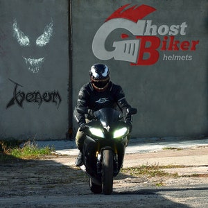 May include: A person wearing a black motorcycle helmet with a Venom design rides a black motorcycle. The motorcycle has a white light on the front. The background is a gray wall with the words "Ghost Biker helmets" in red and white.