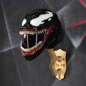 May include: A black and red motorcycle helmet with a Venom character design. The helmet is mounted on a wooden stand.