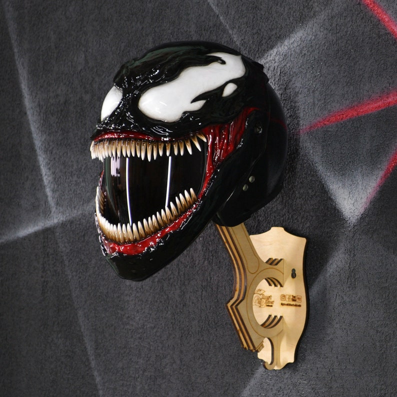 May include: A black and red motorcycle helmet with a Venom character design. The helmet is mounted on a wooden stand.