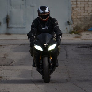 May include: A person wearing a black motorcycle helmet with a white Venom design rides a black motorcycle down a paved road.