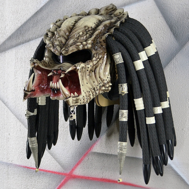 Puede incluir: Un casco de moto blanco y negro con un dise&ntilde;o inspirado en Predator. El casco tiene una boca grande abierta con dientes afilados y una peluca de estilo rastas negra y blanca.