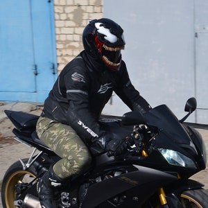 May include: A person wearing a black motorcycle helmet with a Venom design, riding a black motorcycle with a gold rim.