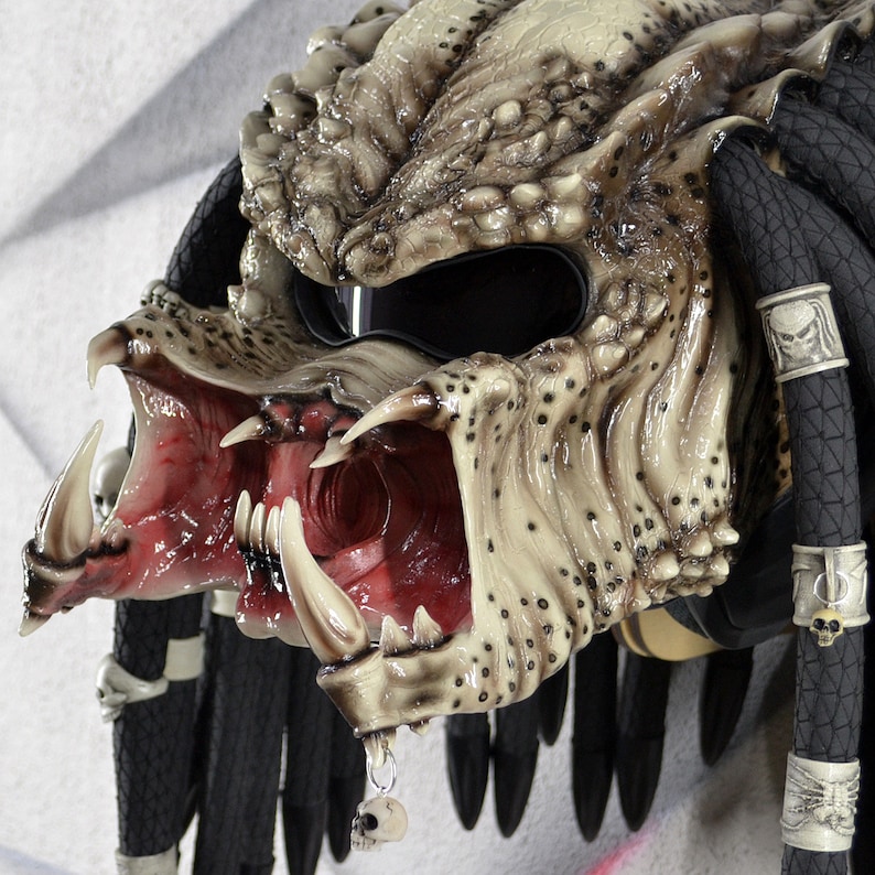 Puede incluir: Un casco de moto con un dise&ntilde;o inspirado en Predator. El casco est&aacute; pintado en un esquema de color beige y marr&oacute;n con una boca roja y dientes afilados. El casco tiene rastas negras con abalorios en forma de calavera.