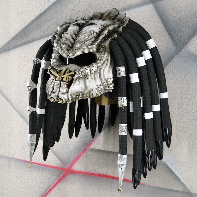 May include: A white motorcycle helmet with a Predator design and black dreadlocks. The helmet has a silver face mask with a black visor and silver accents. The dreadlocks are adorned with silver rings and charms.