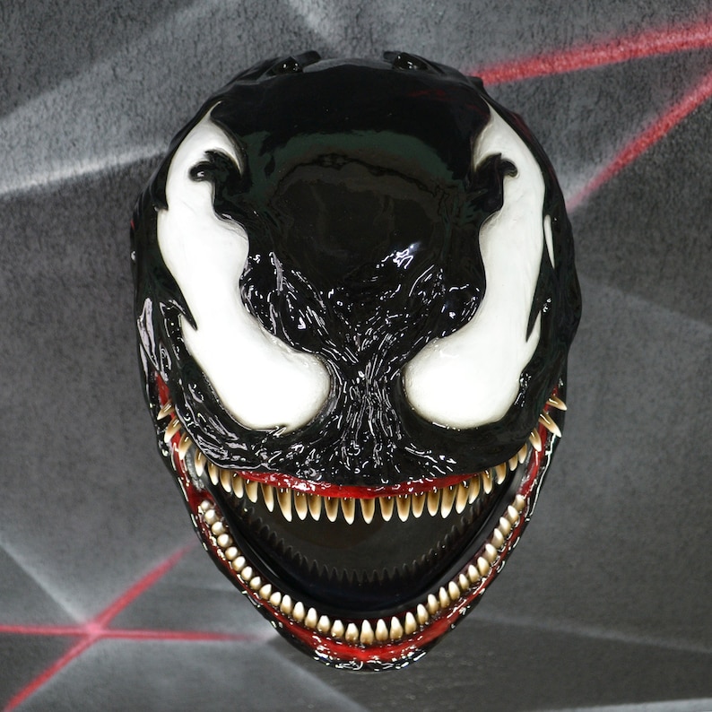 May include: A black and white Venom mask with a large, toothy grin. The mask is made of a hard plastic and has a glossy finish.