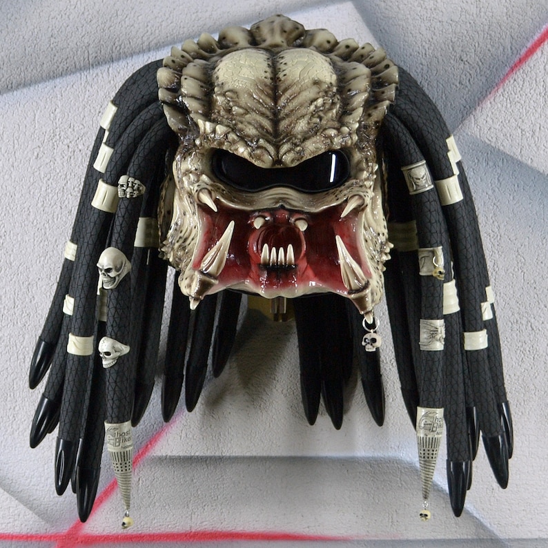Puede incluir: Un casco de moto de estilo Predator blanco y rojo con rastas negras y detalles de calavera.