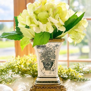 May include: A square ceramic vase with a cream-colored crackle glaze and black floral transferware patterns. The vase sits on a decorative gold-toned metal base with four feet and is filled with a large bouquet of pale green hydrangea flowers.
