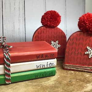 May include: Three wooden blocks stacked on top of each other, painted red, white, and green. The blocks are decorated with the words "warm", "winter", and "wishes". Two red wooden blocks with white pom-poms on top are on the right side of the image. Each block has a white snowflake design.