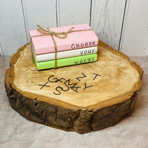 May include: A stack of three small wooden books with painted pink, white, and green covers. The top book has the words "Choose your words" written on it. The books are sitting on a wooden slice with a carved design.
