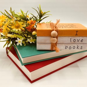 May include: A stack of three books with a yellow, white, and white cover. The top book has the text "I love books" written on it. The books are sitting on top of a green and red book. A bouquet of yellow flowers is in the background.