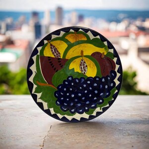 May include: A hand-painted ceramic plate featuring a vibrant fruit design. The plate showcases a colorful arrangement of yellow and green fruits, a cluster of dark blue grapes, and a slice of watermelon. The plate's rim is decorated with a blue and white pattern.