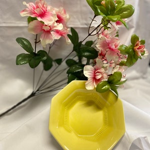May include: A yellow octagonal ceramic dish with a decorative rim. The dish is surrounded by a bouquet of pink and white flowers with green leaves.