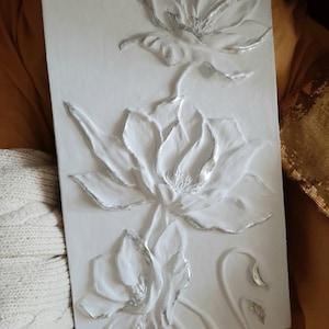 May include: White wall art featuring a 3D relief design of three white flowers with silver accents. The flowers are arranged vertically, with the largest flower at the bottom and the smallest flower at the top.
