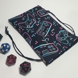 Puede incluir: Una bolsa de cordón negro con un patrón repetido de mandos de videojuegos de color rosa, azul y verde neón. La bolsa está hecha de tela y tiene un cierre de cordón.