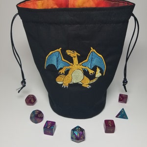 Könnte beinhalten: Ein schwarzer Beutel mit Kordelzug, der vorne einen gestickten orangefarbenen Drachen mit blauen Flügeln zeigt. Das Innere ist mit einem leuchtend orange-roten Stoff gefüttert. Sieben mehrflächige, violette und blaugrüne Würfel liegen im Halbkreis.