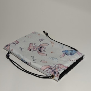May include: A white drawstring bag with a repeating pattern of cartoon characters in pink and blue. The bag has a black drawstring closure and a black interior. The word "Angel" is printed on the bag.