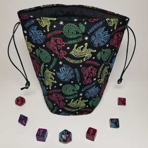 May include: A black fabric dice bag with a drawstring closure. The bag is printed with a repeating pattern of colorful crests and mythical creatures from the Harry Potter universe. The crests include Gryffindor, Hufflepuff, Ravenclaw, and Slytherin. The bag is filled with various colored dice.
