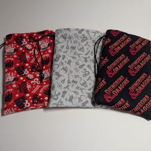 May include: Three drawstring dice bags. One is red with white text and graphics, another is light gray with a pattern, and the third is black with red "Dungeons & Dragons" text. Each bag has a black drawstring.