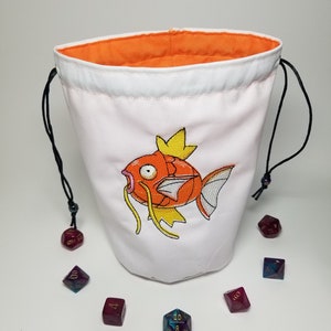 May include: White drawstring bag with an orange lining and an embroidered image of a pink and orange fish with yellow fins. The bag is surrounded by six multi-colored dice.