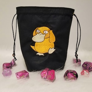 Op de afbeelding: Zwarte trekkoordtas met een geborduurd Psyduck-ontwerp in geel. De tas is omringd door roze en paarse dobbelstenen.