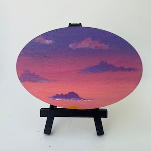 May include: A hand-painted sunset scene on a round wooden canvas. The sky is a gradient of pink and purple with white clouds. A yellow sun is setting behind a purple mountain range.