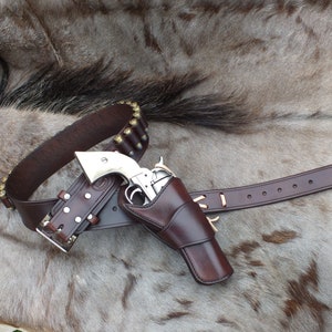 Könnte beinhalten: Ein brauner Leder-Pistolengürtel mit einem silbernen Revolver in einem Holster. Der Gürtel hat eine Patronenschlaufe und eine Schnalle. Der Revolver ist ein einhändiger Sechs-Schuss-Revolver.