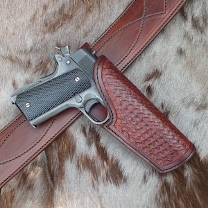 May include: A semi-automatic pistol with a black textured grip and metallic frame rests inside a brown leather holster. The holster features a detailed basketweave pattern and is attached to a matching brown leather belt with stitched diamond accents.