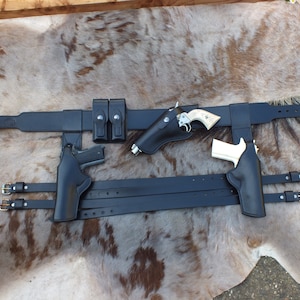 Puede incluir: Un cinturón de pistola de cuero negro con múltiples fundas y bolsas para cargadores. El cinturón se muestra sobre un fondo de piel de vaca blanca y marrón.