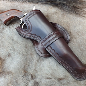 May include: A brown leather holster with intricate stitching and a decorative design. The holster is designed to hold a revolver, which is partially visible in the image.