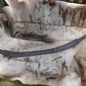 May include: A brown leather gun belt with a row of bullets attached to it. The belt is lying on a white and brown animal hide.
